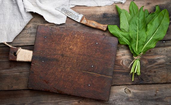 bunch of fresh green sorrel leaves and old brown cutting board on a wooden table, top view