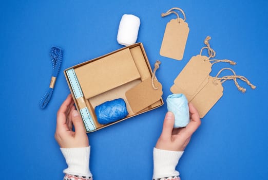 brown cardboard box with a set of festive decor for gift wrapping and two female hands, blue background, top view