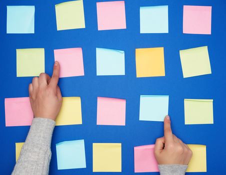 two female hands and a lot of empty paper multi-colored square stickers on a blue background, concept of choosing a strategy with the specified finger