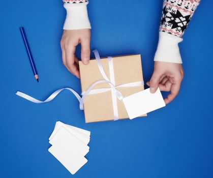 two female hands are holding a cardboard gift square box tied with a white ribbon and empty paper business cards on a dark blue background, top view, classic trend colors