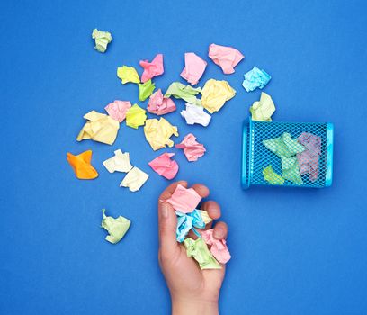 female hand holds a bunch of crumpled multi-colored pieces of paper, next to a metal garbage bin,