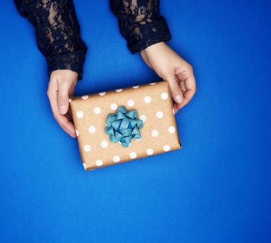 two female hands hold a square brown box with a bow on a blue classic background, top view, festive backdrop