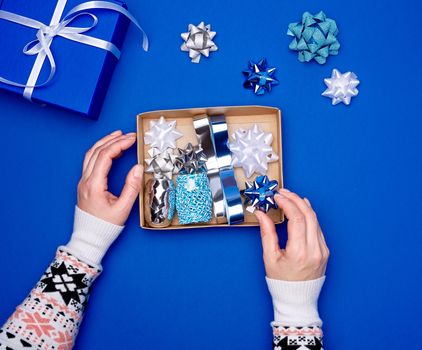 two female hands are holding a brown cardboard box with a set of decor ribbons and bows for wrapping gifts on a dark blue background, top view