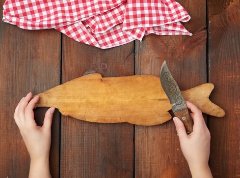 old empty wooden rectangular cutting board and hands hold a sharp knife, top view, brown wooden background 
