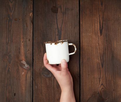 hand holds a white metal cup with rust on a brown wooden background, close up