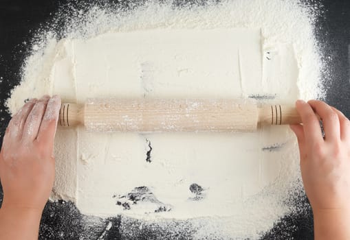 wooden rolling pin in female hands on a black background with white wheat flour, top view
