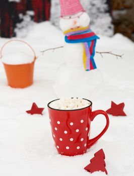 red ceramic mug with hot chocolate and marshmallow, behind a cheerful snowman in the snow