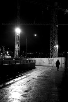 Dark grungy motion blur of unrecognizable woman walking next to a construction site with cranes and sharp lights.