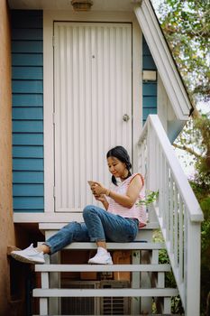 Young asian woman in casual clothes using smartphone and sitting in front of the house for communication and technology concept