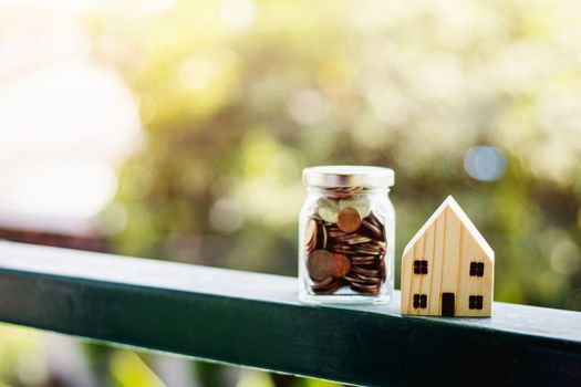 Wooden house model with money coins in the glass jar against blurred natural outdoor background with copy space for business and finance for property concept