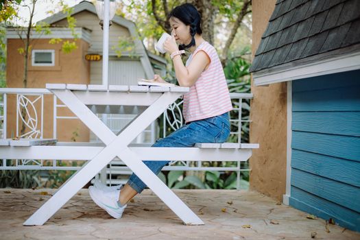 Young asian girl sitting on the white wooden table, drinking a cup of coffee and reading a book at the terraces for relaxation