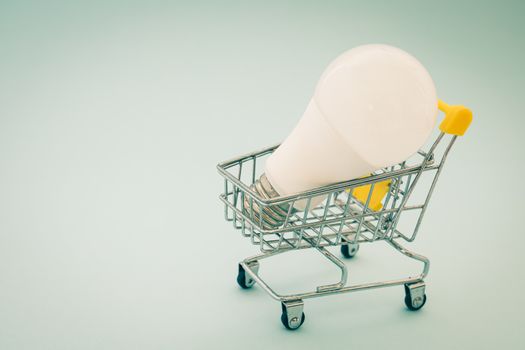 Glowing LED light bulb with mini shopping cart or trolley on blue background for energy savings concept
