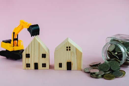 Wooden houses with excavator loader model and money coins in the glass jar on pink background with copy space for business, property finance and construction concept