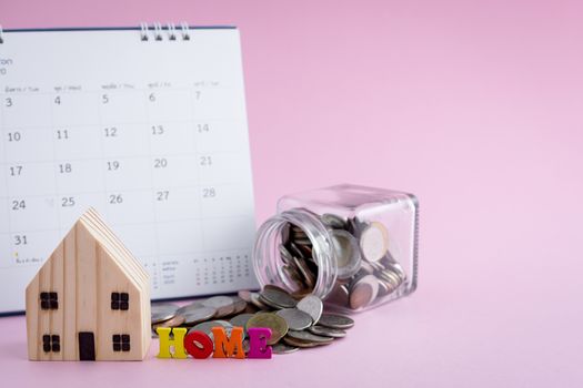 Wooden house model with HOME alphabet, money coins in the glass jar and calendar on pink background for housing business and planning concept
