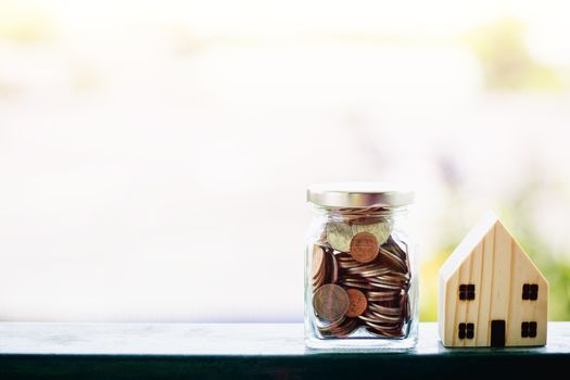 Wooden house model with money coins in the glass jar against blurred natural outdoor background with copy space for business and finance for property concept