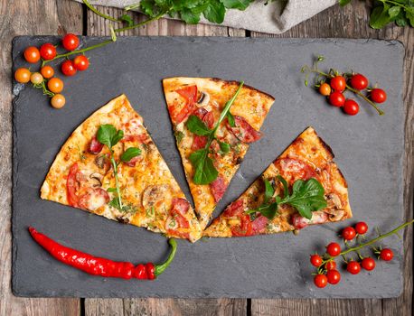 triangular piece of baked pizza with mushrooms, smoked sausages, tomatoes and cheese, black background, flat lay