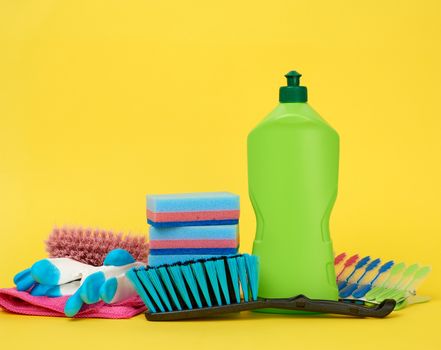 rubber gloves for cleaning, multi-colored sponges, brushes and cleaning fluid in a green plastic bottle on a yellow background