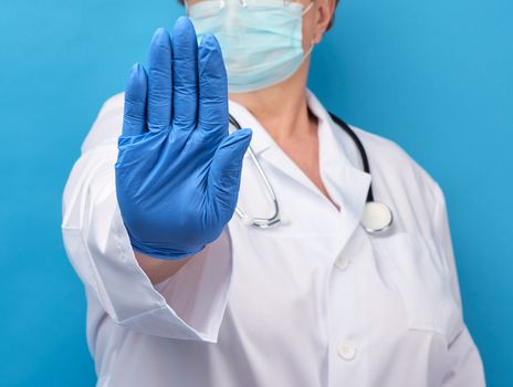 female doctor in white medical coat and blue latex gloves shows stop gesture, concept of keeping a safe distance, do not panic