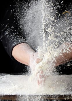 chef in black uniform sprinkles white wheat flour in different directions, product scatters dust, black background