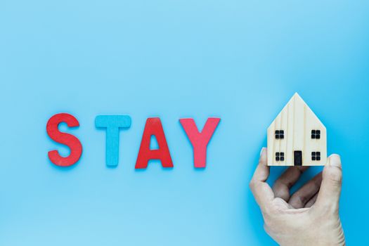 Hand holding wooden house model with STAY text on blue background for self-quarantine, staying home and social distancing in coronavirus or Covid-2019 situation concept