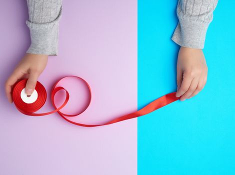 two female hands holding a bobbin with a red silk ribbon on a colored background, top view