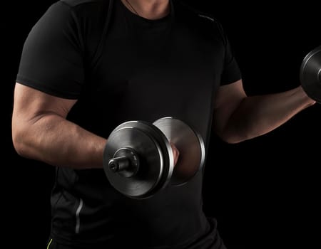 man in black clothes holds steel dumbbells in his hands, his muscles are tense, low key, sports background