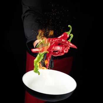 chef in a black uniform holds a round pan and throws up red and green whole chili peppers in a burning fire, cooking spices, low key