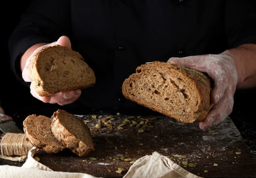 chef in black uniform keeps cut off a piece of bread baked from rye flour and pumpkin seeds, black background