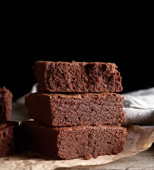 stack of baked square pieces of chocolate brownie cake on brown wooden cutting board, black background