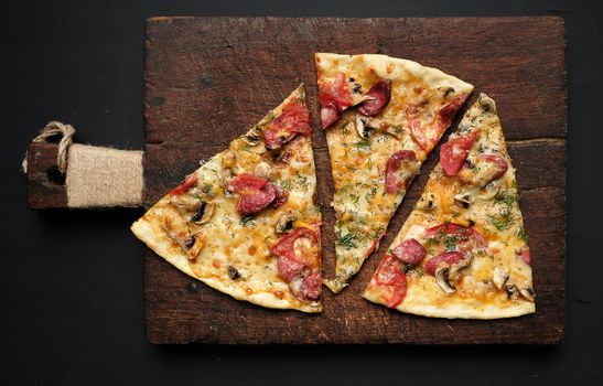 baked pizza with smoked sausages, mushrooms, tomatoes, cheese and dill, sliced food on a brown wooden board, close up