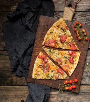 baked pizza with smoked sausages, mushrooms, tomatoes, cheese and dill, sliced food on a brown wooden board, close up