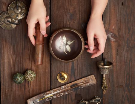 female hands hold a copper Tibetan singing bowl and a wooden stick, on the wooden table are items for alternative medicine, sound massage and meditation