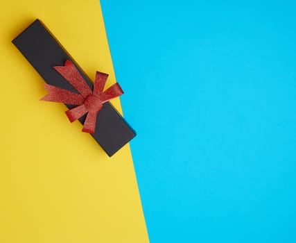 closed black paper cardboard box with a red bow on a yellow-blue background, concept of surprise, place for text
