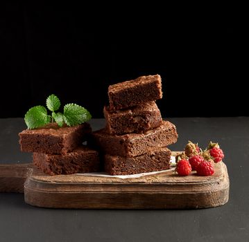 stack of baked square pieces of chocolate brownie cake on brown wooden cutting board, black background