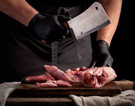 chef in black latex gloves holds a big knife and cuts into pieces raw rabbit meat on a brown wooden cutting board, cooking on a dark background