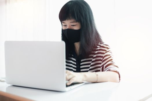 Woman wearing a black face mask, using laptop and working at home for business, self-quarantine, staying home and social distancing in coronavirus or Covid-2019 outbreak situation concept