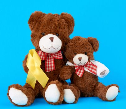 brown teddy bear sits and holds in his paw a yellow silk ribbon on a blue background, concept of the fight against childhood cancer. problem of suicides and their prevention