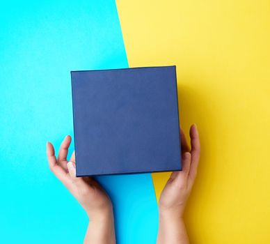 two female hands hold a closed dark blue paper cardboard box on a yellow-blue background, concept of surprise, top view