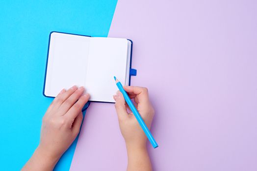 open notebook and wooden blue pencil in a female right hand on a colored background, top view, process of writing information on paper