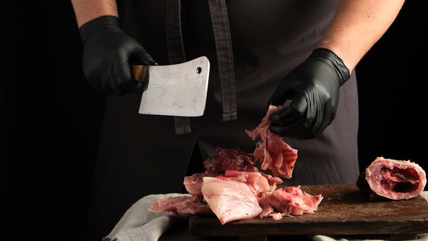 chef in black latex gloves holds a big knife and cuts into pieces raw rabbit meat on a brown wooden cutting board, cooking on a dark background