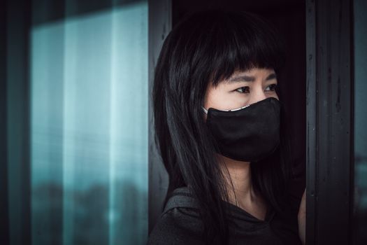 Asian woman wearing a black face mask, standing absent-minded at the door and staying home for self-quarantine and social distancing in coronavirus or Covid-2019 outbreak situation concept