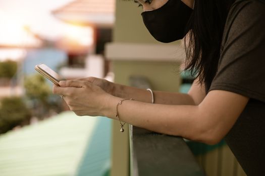 Asian woman wearing a black face mask, using smartphone at the terrace and staying home for self-quarantine and social distancing in coronavirus or Covid-2019 outbreak situation concept