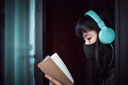 Asian woman wearing a black face mask and headphones, reading a book and staying home for self-quarantine and social distancing in coronavirus or Covid-2019 outbreak situation concept