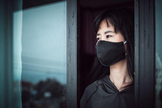 Asian woman wearing a black face mask, standing absent-minded at the door and staying home for self-quarantine and social distancing in coronavirus or Covid-2019 outbreak situation concept