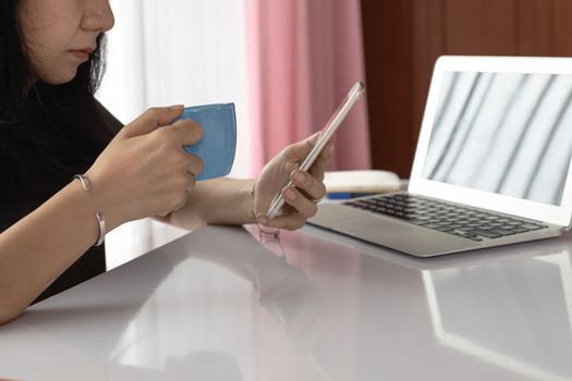 Woman drinking and using smartphone with computer laptop and working at home for business, self-quarantine, staying home and social distancing in coronavirus or Covid-2019 outbreak situation concept