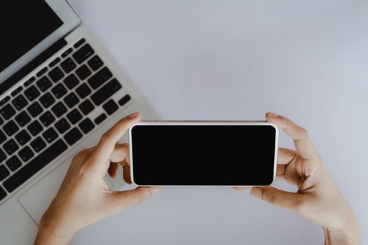 Top view of hand holding smartphone with blank black screen and computer laptop on white office desk for business, communication and media technology concept