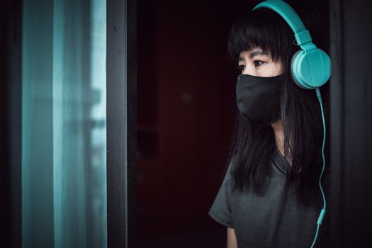 Asian woman wearing a black face mask and headphones, standing absent-minded at the door and staying home for self-quarantine and social distancing in coronavirus or Covid-2019 outbreak situation