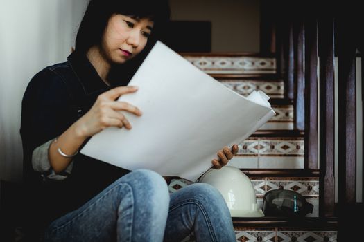 Asian woman looking architectural plan, sitting on the stairs and working at home with safety helmet for building inspection concept