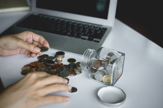 Woman's hand saving and counting money coins in the glass jar with computer laptop for planning, business and finance concept