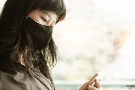 Asian woman wearing a black face mask, using smartphone at the door and staying home for self-quarantine and social distancing in coronavirus or Covid-2019 outbreak situation concept